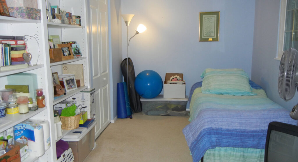 A cluttered bedroom with a bed, bookshelf, exercise equipment, and a wall-mounted fan.