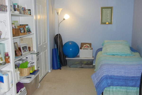 A cluttered bedroom with a bed, bookshelf, exercise equipment, and a wall-mounted fan.