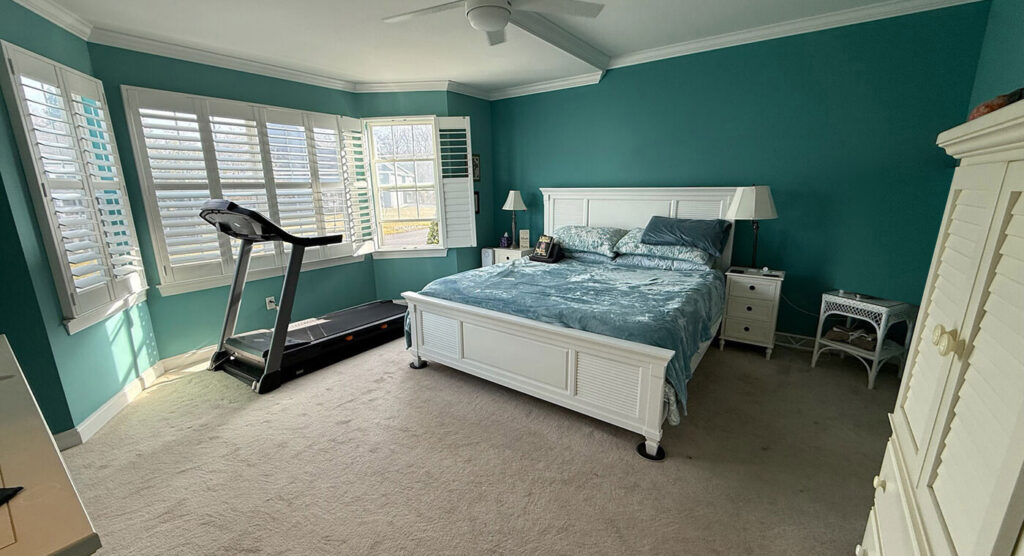A bedroom with teal walls, a white bed, a treadmill by the window, and a white wardrobe, illuminated by natural light.