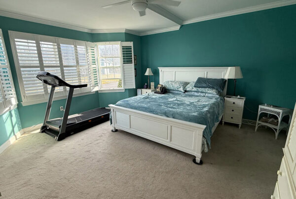 A bedroom with teal walls, a white bed, a treadmill by the window, and a white wardrobe, illuminated by natural light.