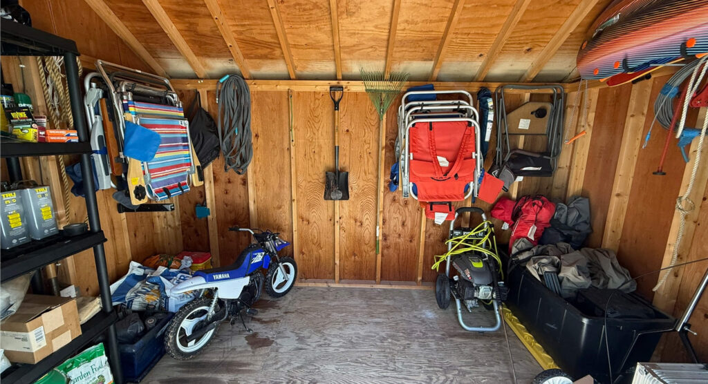 A wooden garage interior with tools, a toy bike, folding chairs, a snowblower, and various outdoor gear hanging and stored