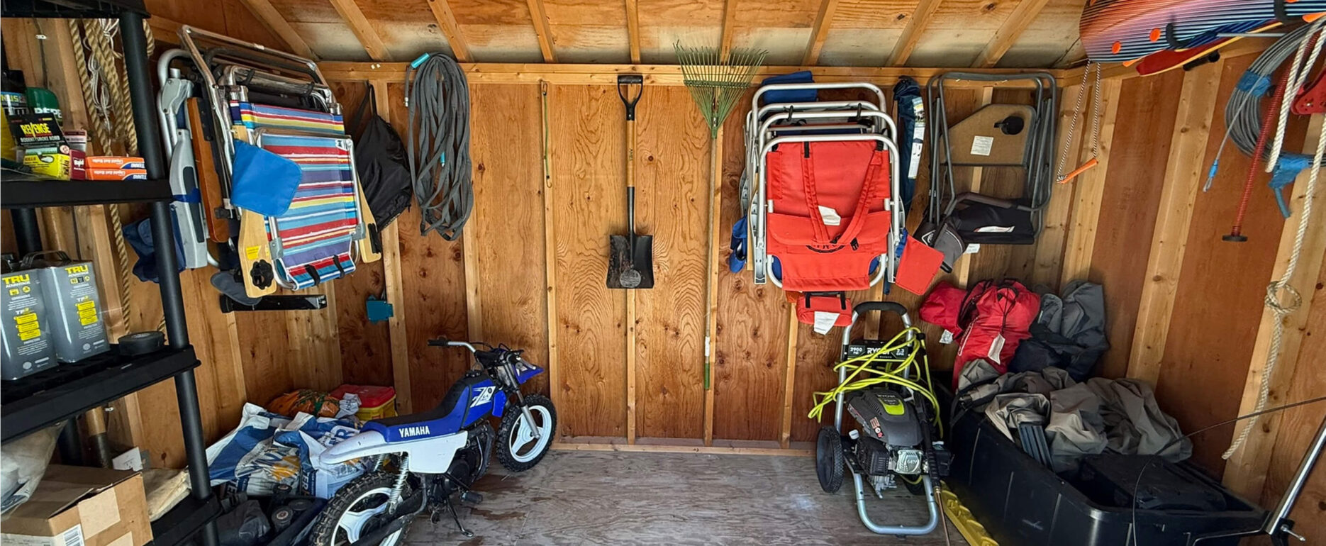 A wooden garage interior with tools, a toy bike, folding chairs, a snowblower, and various outdoor gear hanging and stored