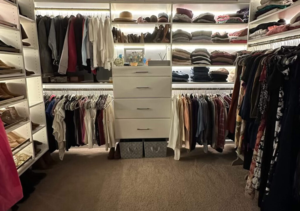 A walk-in closet with shelves of folded clothes, hanging shirts and pants, and a central white dresser with decorative items