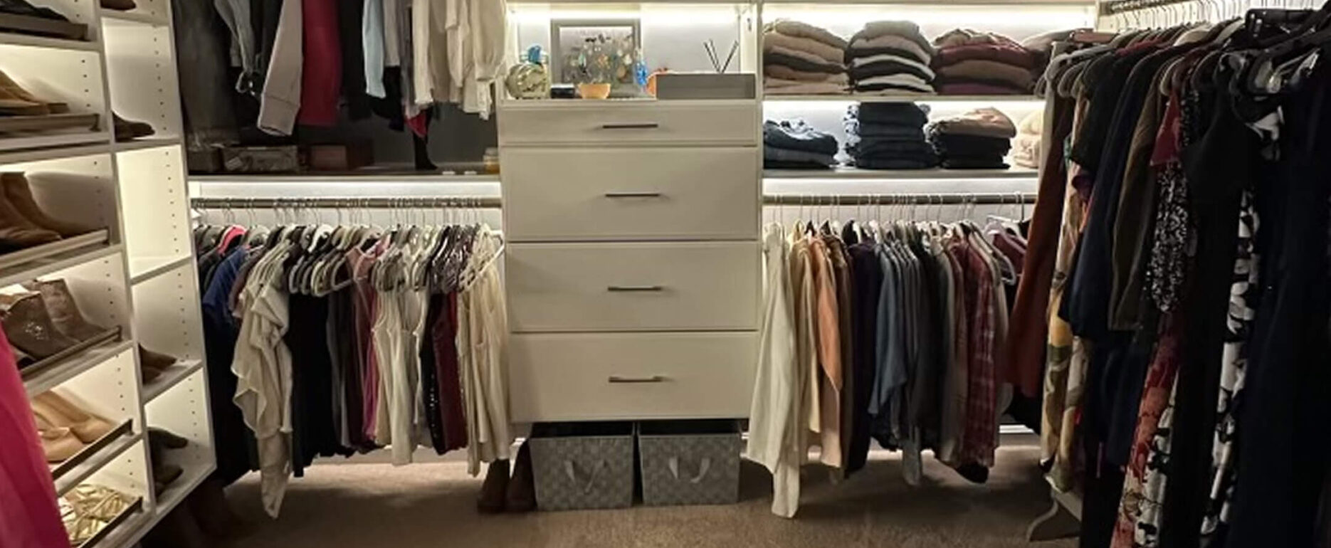 A walk-in closet with shelves of folded clothes, hanging shirts and pants, and a central white dresser with decorative items