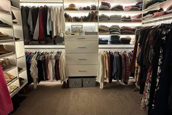 A walk-in closet with shelves of folded clothes, hanging shirts and pants, and a central white dresser with decorative items